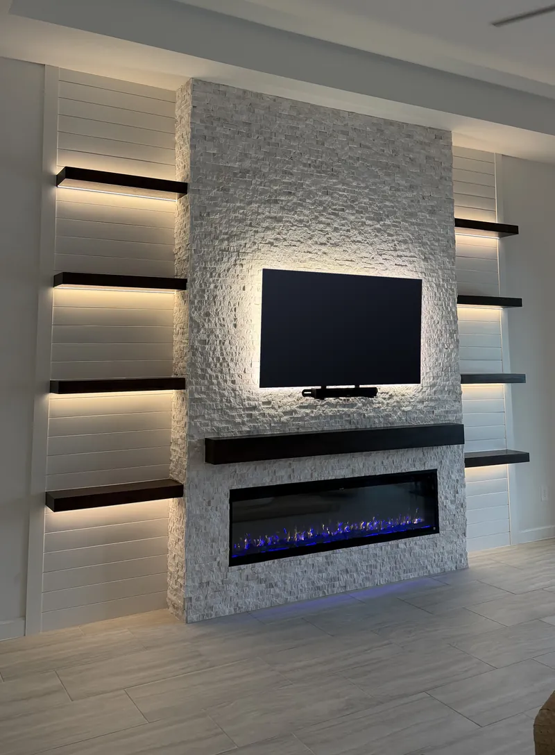 White stacked stone wall rising to the ceiling with a backlit TV, a soundbar, an espresso-tone mantel and a linear fireplace, between white shiplap and four lit floating shelves on each side