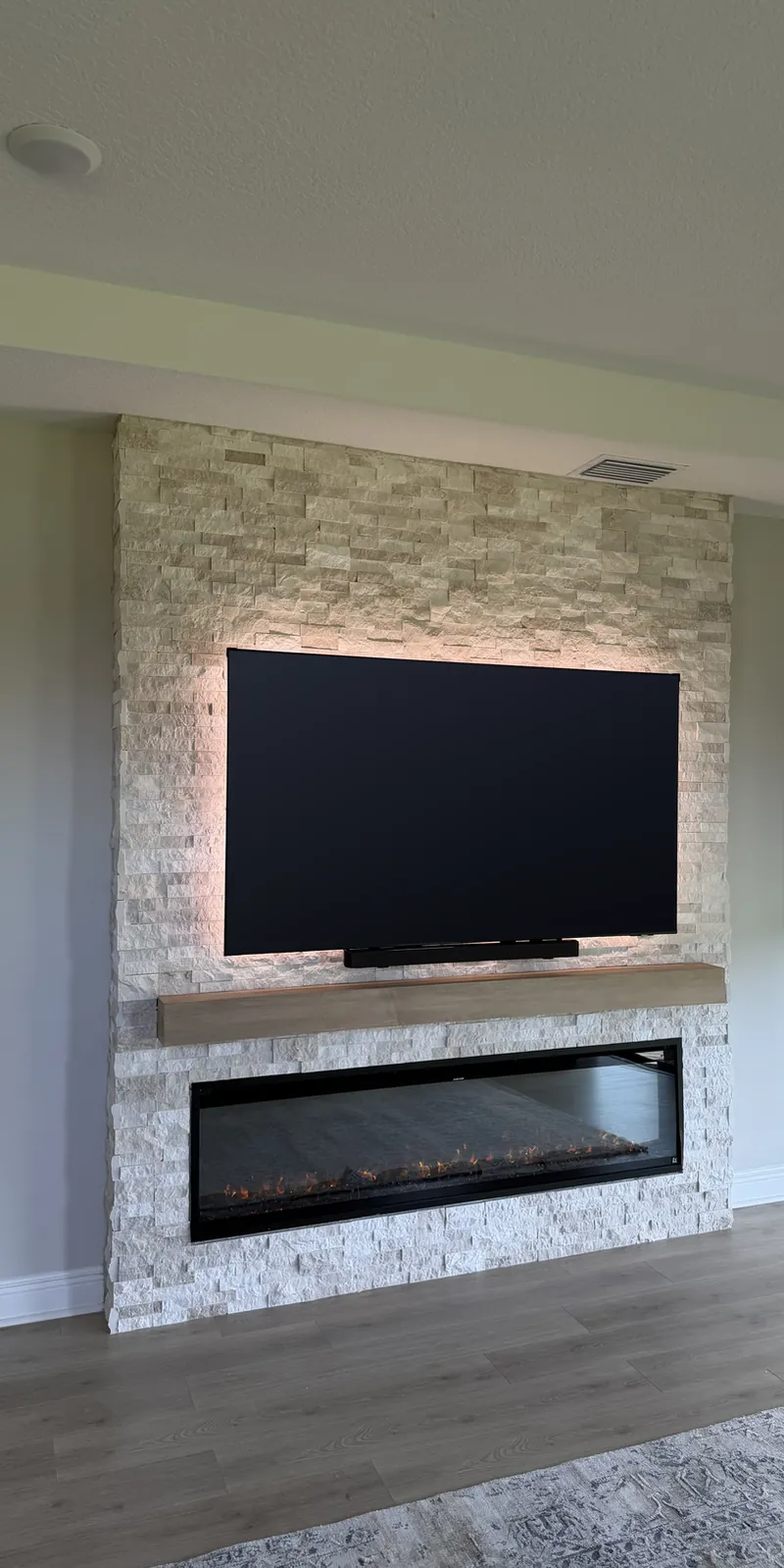 Stacked stone column fireplace wall with a backlit TV, a light wood-tone mantel and a long linear fireplace