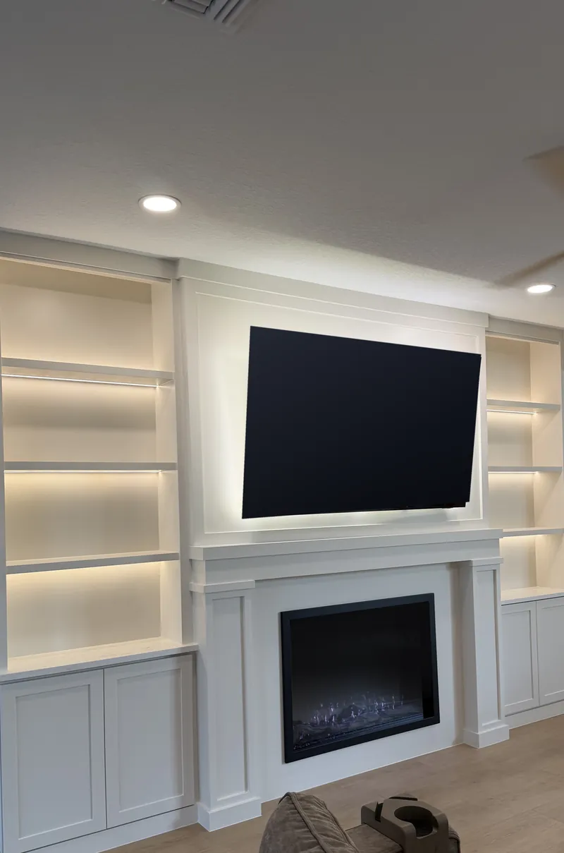 All-white built-in wall with the TV above a lit fireplace in a classic surround, between tall lit towers