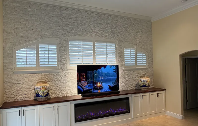 Stacked stone wall around three shuttered windows over a long white credenza with a linear fireplace