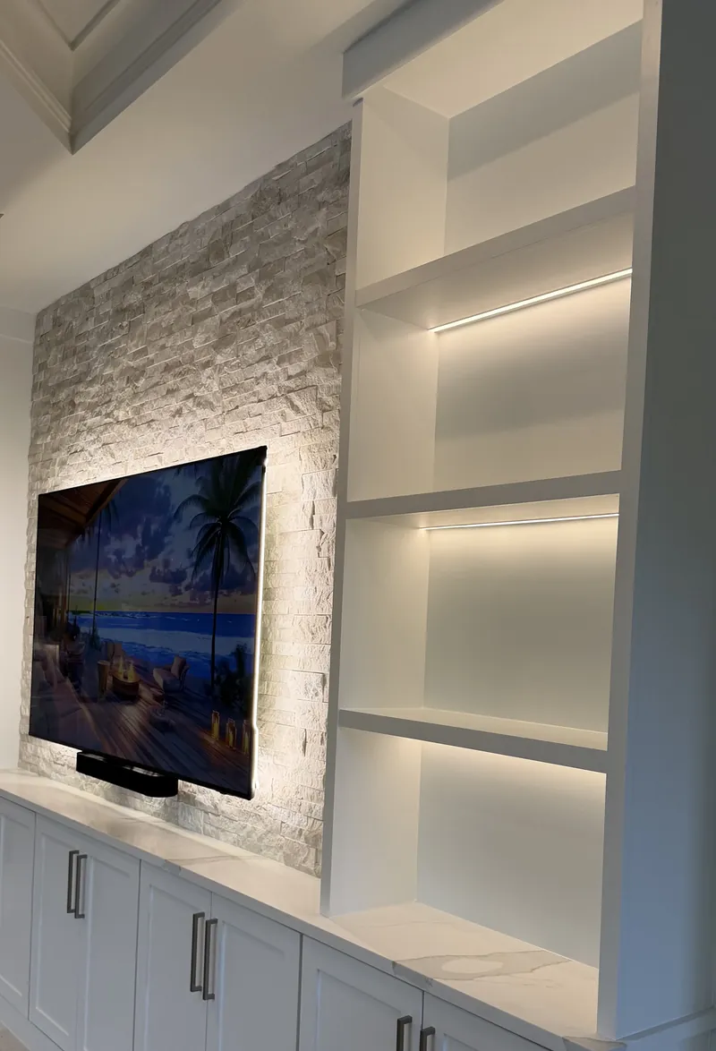 Cream stacked stone behind a backlit TV, with open white towers lit under each shelf over Shaker cabinets