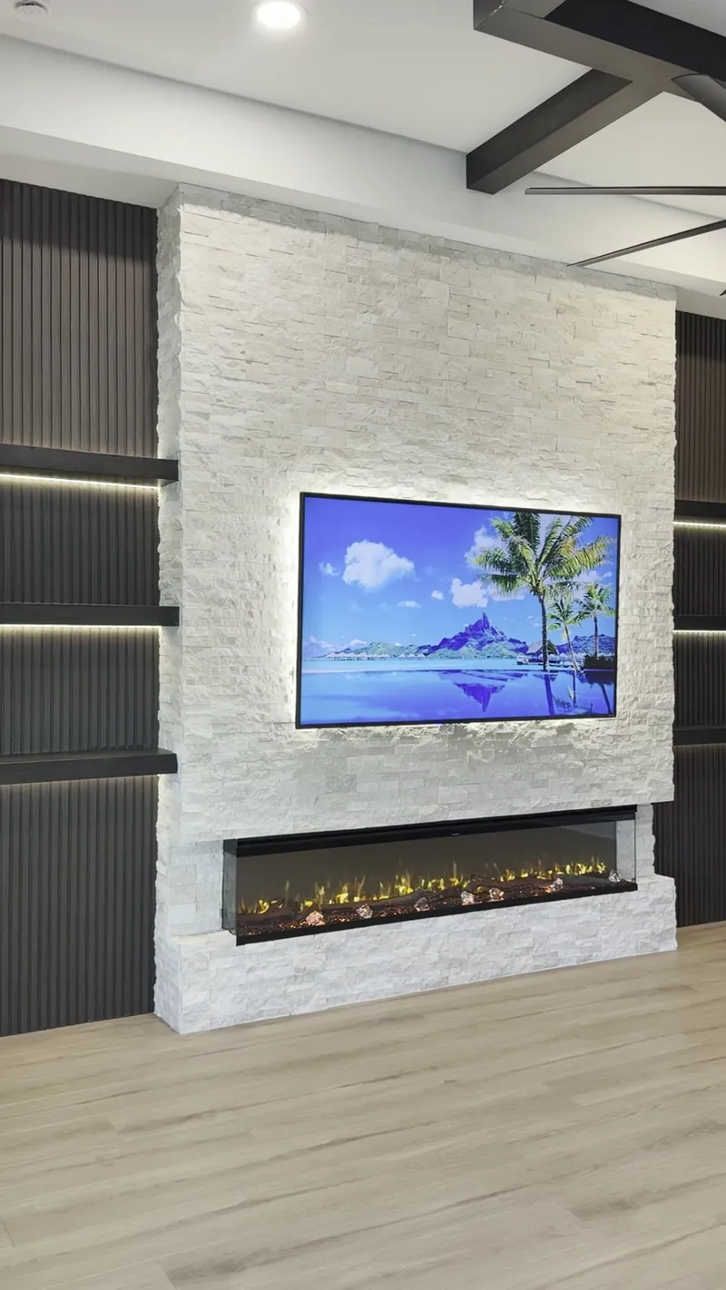 The Midnight Fluted Stone Wall: white stacked stone with a backlit TV over a three-sided fireplace, between dark fluted panels with lit floating shelves