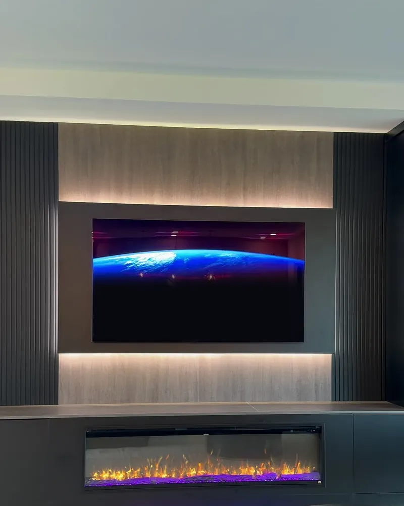 TV on a dark panel between two lit light wood-tone bands, dark slats on each side and a linear fireplace below